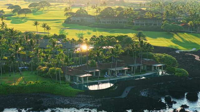 Drone aerial view of luxury oceanfront villas with infinity pool palm trees and green golf course beside black lava rock coastline at golden sunrise on Big Island Hawaii.