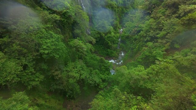FPV aerial view over lush rainforest gorge in Costa Rica with cascading waterfall mist rising above dense green canopy and rocky river flowing through steep tropical jungle valley.