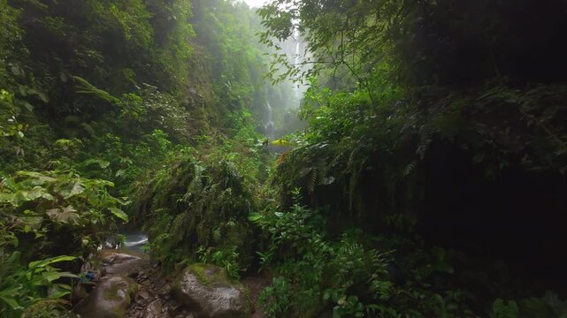 FPV drone flight through misty rainforest canyon in Costa Rica revealing tall waterfall lush ferns mossy boulders and turquoise river rapids with hikers exploring wild tropical jungle gorge.