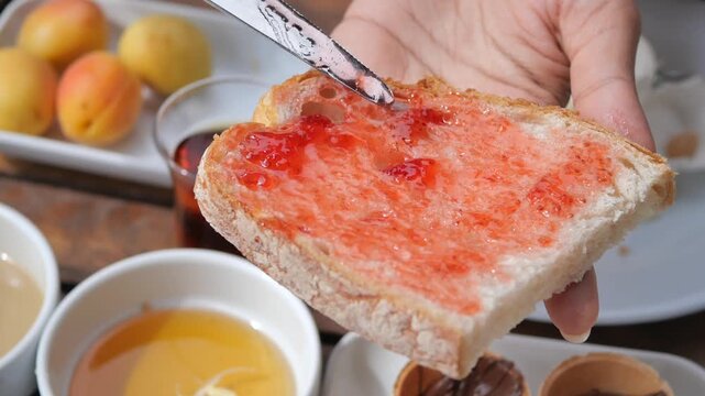 Close up hands spreading strawberry jam on slice of bread, delicious breakfast spread on rustic wooden table, healthy morning meal preparation