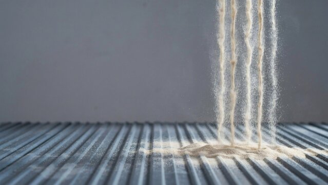 Pale sand pouring in thin sheets over a ridged steel plate in studio light