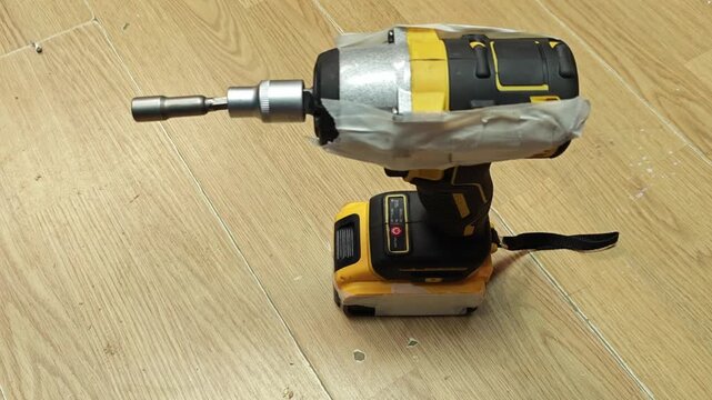 Impact driver product demo on wood floor. Cordless impact driver with metal bit extension stands alone for close inspection, showing taped housing and battery pack in simple workshop style setup.