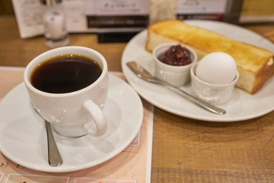 A white cup of black coffee rests on a wooden table with blurred buttered toast a boiled egg and sweet red bean paste in the background Ideal for food blogs and cafe menu marketing