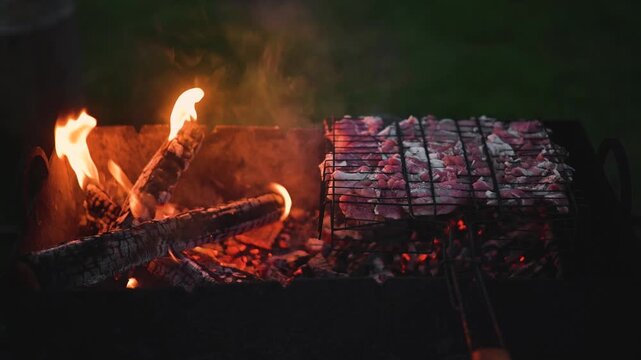 Meat is being cooked on a grill over an open flame, with visible flames and smoke rising. The setting appears to be outdoors, showcasing the grilling process