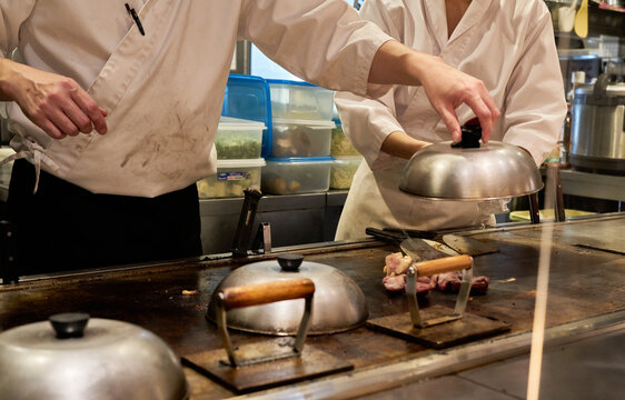 A Japanese chef in a white uniform lifts a metal dome over cooking meat on a hot iron griddle in a commercial kitchen Ideal for restaurant management articles and culinary arts editorials