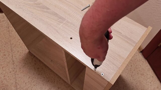 Wood cabinet assembly is underway while hand uses electric screwdriver to fix screws in place. Flat pack parts rest on floor, showing careful, focused joinery and practical home furniture setup.