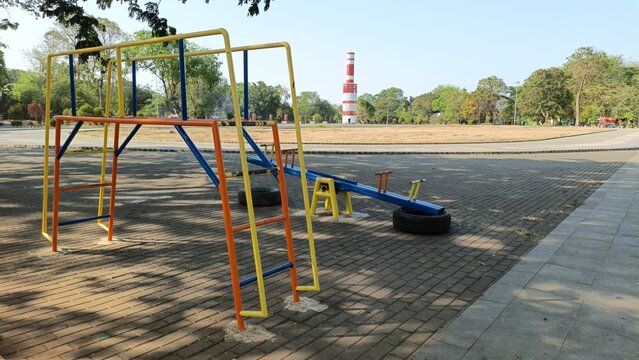 A quiet outdoor public park featuring colorful metal playground equipment, including a climbing frame and seesaw on paved ground, with lush trees and a distant red-and-white tower under a clear daytim