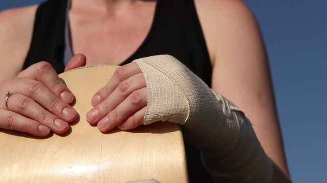 Skateboarder sits with bandaged hand and skateboard against blue sky. Skateboarding injury, first aid, immobilization of injured hand, break in skating. Skateboard in girl's hands