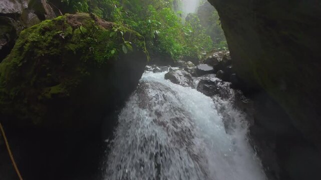 FPV drone flies low over rushing jungle river toward hidden misty waterfall in Costa Rica rainforest with hikers standing on wet mossy boulders surrounded by lush tropical greenery.