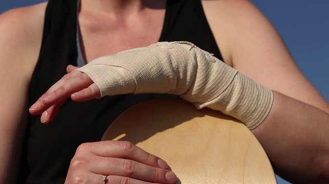 Skateboarder sits with bandaged hand and skateboard against blue sky. Skateboarding injury, first aid, immobilization of injured hand, break in skating. Skateboard in girl's hands