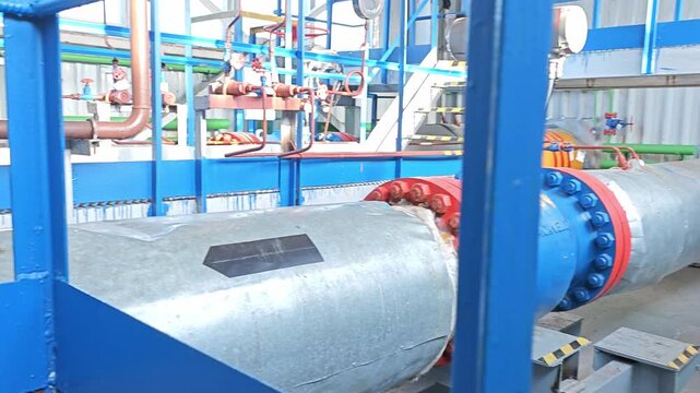 Industrial infrastructure network stands inside plant room, with oversized insulated pipes, blue valve housings, and red flanges. Bright metal pipework creates controlled, technical utility setting.