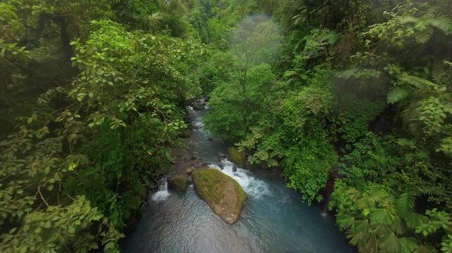 FPV drone flies low over a rocky jungle river in Costa Rica racing between mossy boulders and dense green rainforest foliage toward a misty waterfall in a steep tropical gorge.