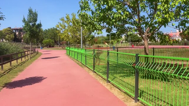 Red rubberized jogging track bordered by timber handrail and bright green steel mesh fencing in modern urban public park during clear summer daylight. High quality 4k footage