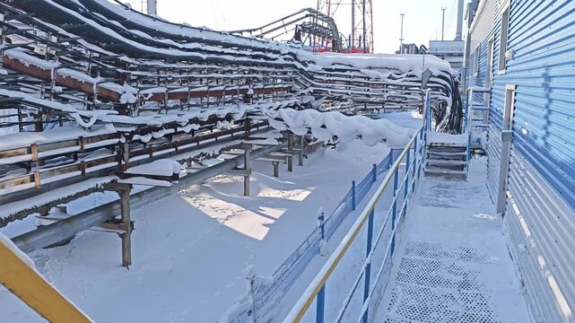 Snow covered pipeline runs through industrial walkway in winter. Blue metal railings and pipe racks line narrow service path, showing frozen outdoor infrastructure at quiet processing facility.