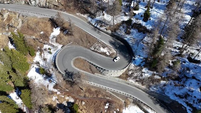 white car rounds tight hairpin bend stone retaining wall guardrail sparse larch snow patches malojaschlange switchback maloja pass drone footage graubunden switzerland