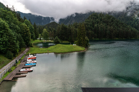 Colorful pedal boats are moored at a wooden dock on Fernsteinsee in Tyrol, Austria. Emerald lake water, grassy shore, evergreen forest and cloud covered Alpine mountains create a scenic recreation and
