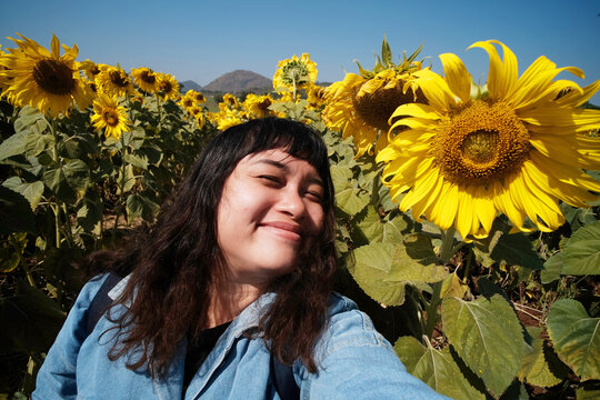 Smiling Asian female traveler holding camera and selfie with yellow sunflower field and blue sky. happiness with a sense of freedom in the nature.