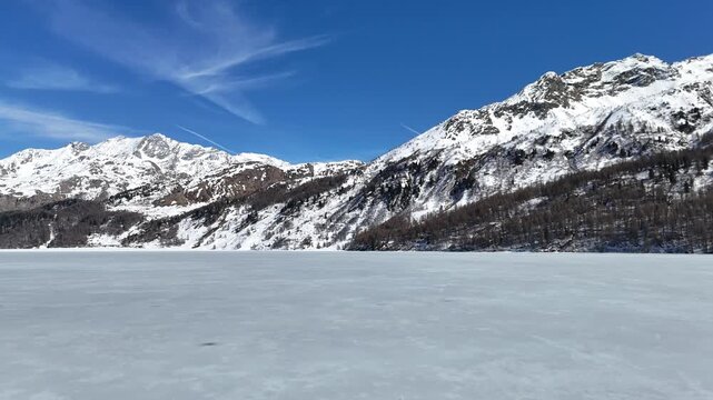 frozen lake surface stretches toward snow capped mountain ridge forested shoreline clear blue sky wispy clouds silsersee lake sils engadin valley graubunden switzerland swiss alps winter