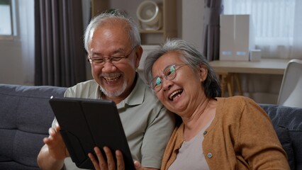 Joyful senior Asian couple laugh together while looking at a tablet at home, portraying positive...