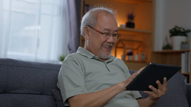 Contented senior Asian man wearing eyeglasses sitting on couch, using mobile application on digital tablet with satisfied smile in warm lighted living room.