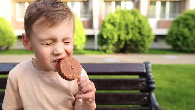 Happy boy sits on hotel bench and bites new chocolate ice cream. Round dessert has rich chocolate coating and abundant sprinkles creating indulgent summer treat.