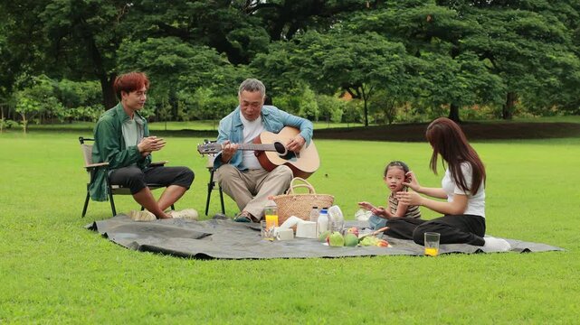 GrandFather playing acoustic guitar for child during picnic. Asian family enjoy outdoor picnic with food and drinks in park sunny day, outdoor activitie with daughter father, mother grandfather