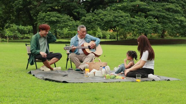 GrandFather playing acoustic guitar for child during picnic. Asian family enjoy outdoor picnic with food and drinks in park sunny day, outdoor activitie with daughter father, mother grandfather