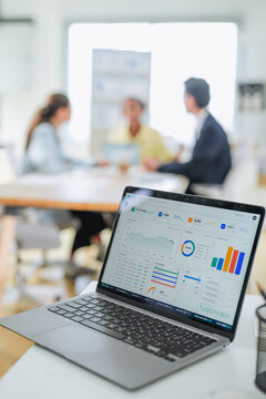 Close-up of a data dashboard on a laptop screen showing sales metrics and performance charts, with a business meeting unfolding softly out of focus behind it.