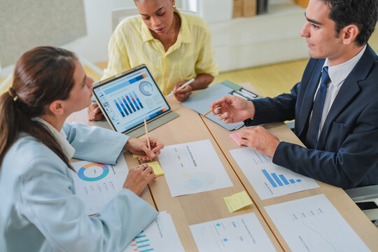 Close-up of business professionals collaborating over performance data, gesturing toward a pie chart on a tablet surrounded by printed reports and sticky notes.