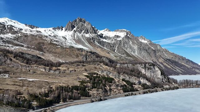 mountain road winds along frozen lake sils shoreline engadin valley switzerland alpine hamlet nestled larch forest slope snowcapped granite peaks winter drone aerial flight view