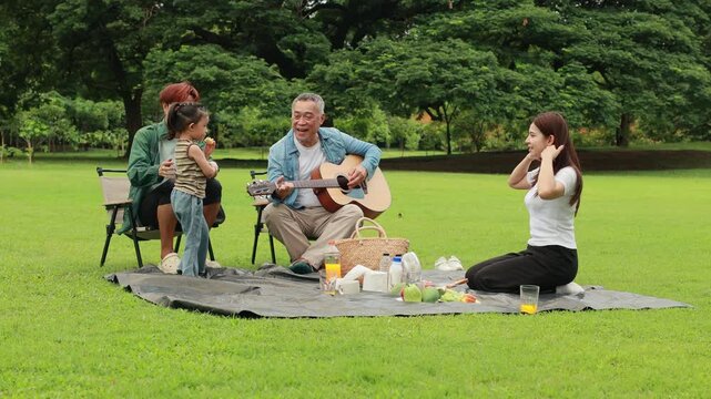 GrandFather playing acoustic guitar for child during picnic. Asian family enjoy outdoor picnic with food and drinks in park sunny day, outdoor activitie with daughter father, mother grandfather