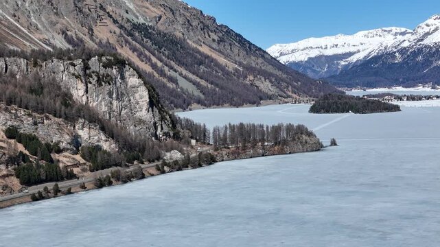 silsersee frozen lake valley wooded island distant alpine village engadin switzerland mountain range granite peaks larch forest winter drone flyover panoramic aerial view