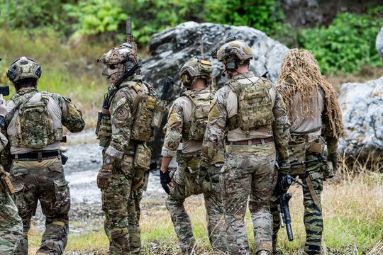 A group of soldiers is gathered in a forested area. They wear tactical gear and appear to be engaged in a training exercise. The scene shows rugged terrain and natural surroundings.