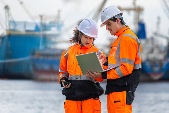 Engineers team in safety gear and hard hats using laptop at port cargo yard, professional logistics operators collaborating and working on computer at shipping terminal