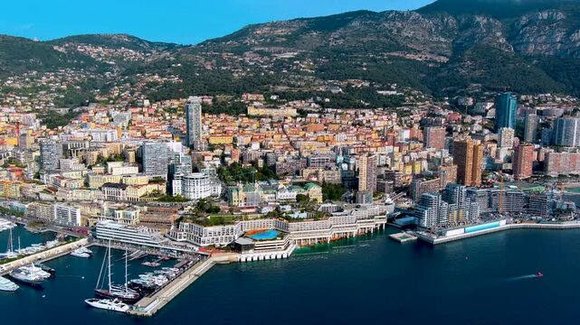 Aerial drone view of Monte Carlo in Monaco with the casino and its green roofs, terraced seafront complexes, Port Hercule marina with yachts, high-rise towers and the mountains behind.