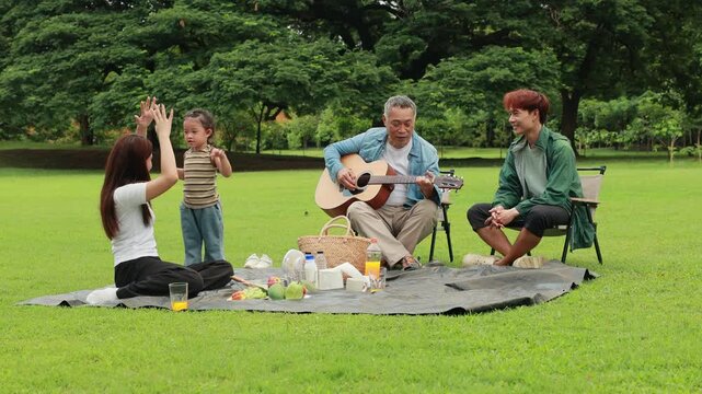 GrandFather playing acoustic guitar for child during picnic. Asian family enjoy outdoor picnic with food and drinks in park sunny day, outdoor activitie with daughter father, mother grandfather