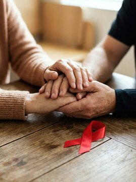 Two people holding hands together on a wooden table with a red awareness ribbon for World AIDS Day. Symbol of compassion, medical support, solidarity, and empathy for patients in need.