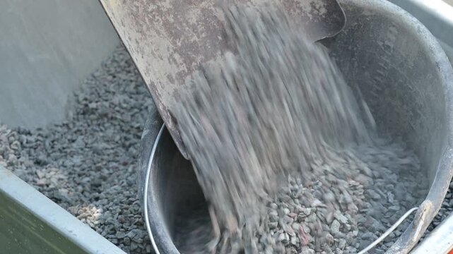Shoveling crushed stone from a wheelbarrow into a bucket on a construction site. Close-up of gray gravel and dust inside a metal wheelbarrow and an old bucket, material preparation