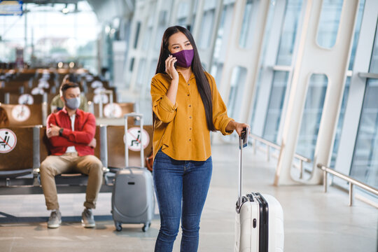 A woman in a face mask talks on her phone while standing with her suitcase in a modern airport waiting area