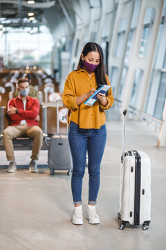 A woman stands in an airport terminal holding her phone and travel documents while a man sits in the background, both wearing face masks