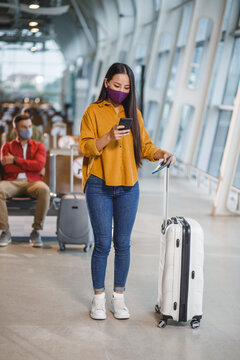 A young woman wearing a purple face mask checks her phone while standing with her suitcase and passport in a modern airport terminal