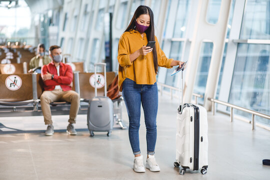 A young woman wearing a face mask stands in an airport terminal, looking at her smartphone while holding her passport and boarding pass near her suitcase