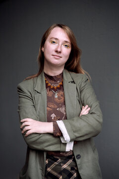 a serious but confident young businesswoman standing with crossed arms against a plain dark grey background. She is wearing a professional olive-green blazer over a pattern blouse, looking directly at