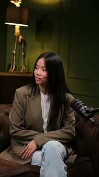 Confident Asian woman sitting on a couch and speaking into a microphone during a podcast recording