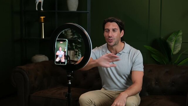 Young man vlogging with his smartphone and a ring light, talking to his online audience at home