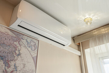 White wall-mounted split air conditioner sits below ceiling, paired with vintage world map, sheer curtains, decorative cornice, crystal ceiling light, neutral beige walls © evgeniykleymenov