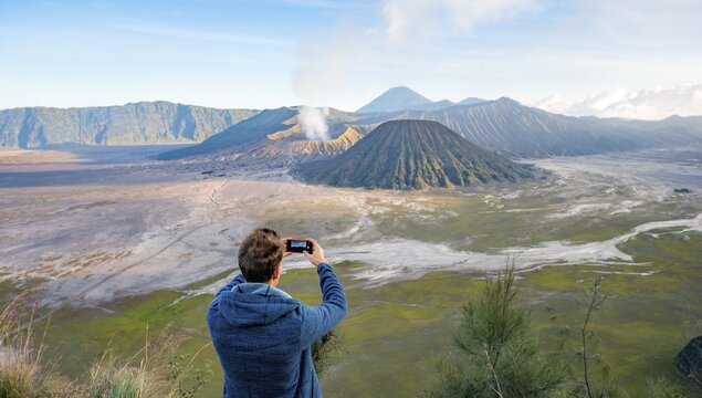 Young man takes photo with iPhone, volcanic landscape, view in Tengger Caldera, smoking volcano Gunung Bromo, in front Mt. Batok, in the back Mt. Kursi, Mt. Gunung Semeru, National Park Bromo-Tengger-Semeru, Java, Indonesia