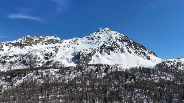 sharp snowcapped summit dominates skyline above dense forested ridge clear blue sky engadin valley switzerland swiss alps graubunden winter drone aerial mountain view