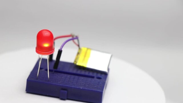 Small breadboard with red LED glowing connected to battery power through a resistor rotating to show the details of the circuit