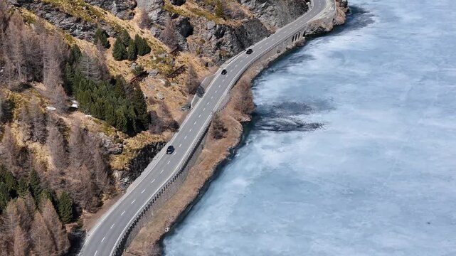 cliffside road winds along frozen silsersee shoreline cars travel past guardrail rocky slope thin ice patch visible on lake surface engadin valley switzerland drone aerial view